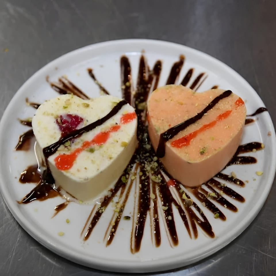 Two heart-shaped Indian desserts, one white and one orange, drizzled with chocolate and red sauce.