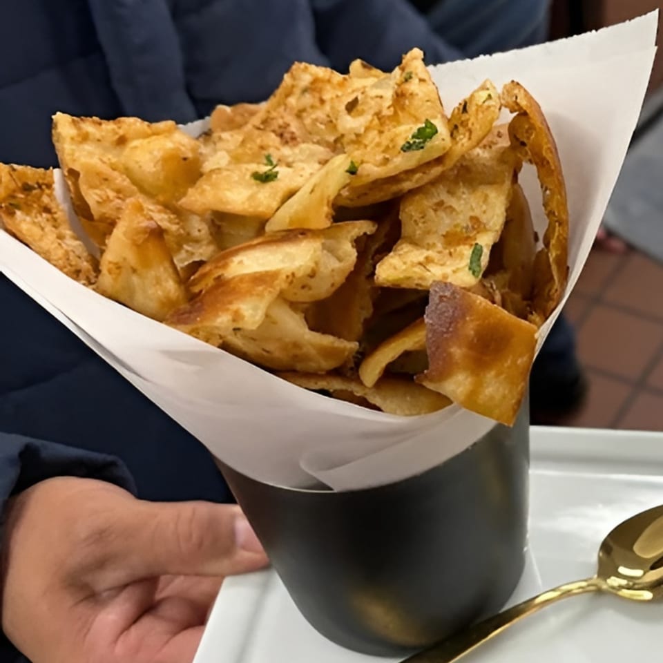 A hand holds a black cup filled with crispy, golden-brown Indian flatbread pieces, garnished with herbs.