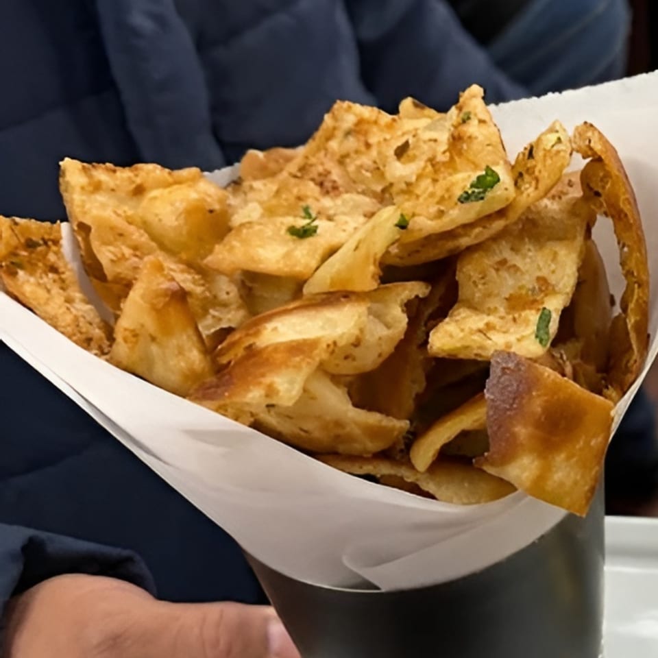 Crispy, golden Indian flatbread pieces, garnished with herbs, served in a white paper cone.