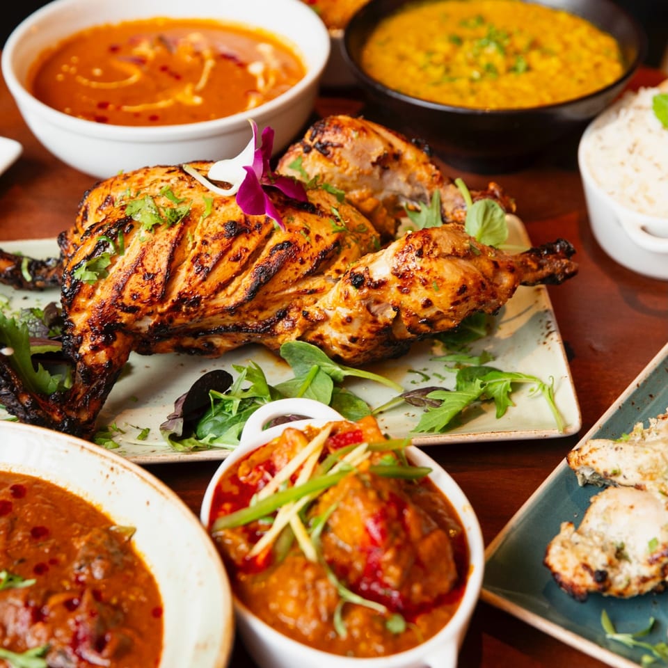 A whole grilled Tandoori chicken, garnished with cilantro and an orchid, surrounded by Indian curries and rice.